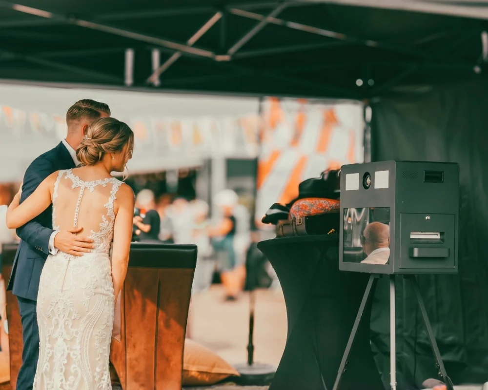 Bruidspaar staat bij een photobooth op hun bruiloft, met feestgasten en decoratie op de achtergrond.